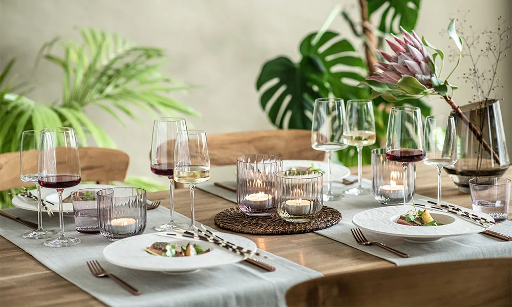 Elegantes, gedecktes Tischarrangement mit Weingläsern, Tellern und Kerzen, umgeben von üppigen grünen Pflanzen.