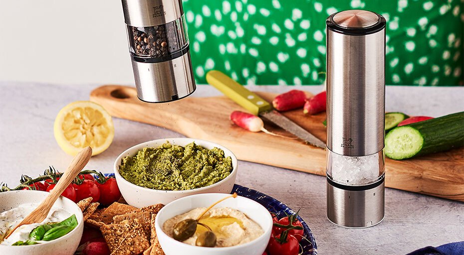 Zwei elektrische Gewürzmühlen der Serie Elis von Peugeot und Dipschalen mit Gemüse auf einem Tisch. Hintergrund: Schneidebrett mit Radieschen.