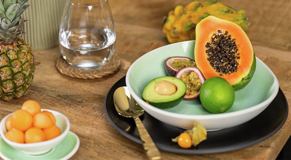 Teller der Serie Junto Jungle von Rosenthal mit exotischen Früchten: Papaya, Avocado, Passionsfrucht, Limette; daneben Melonenkugeln und Physalis auf Holztisch.