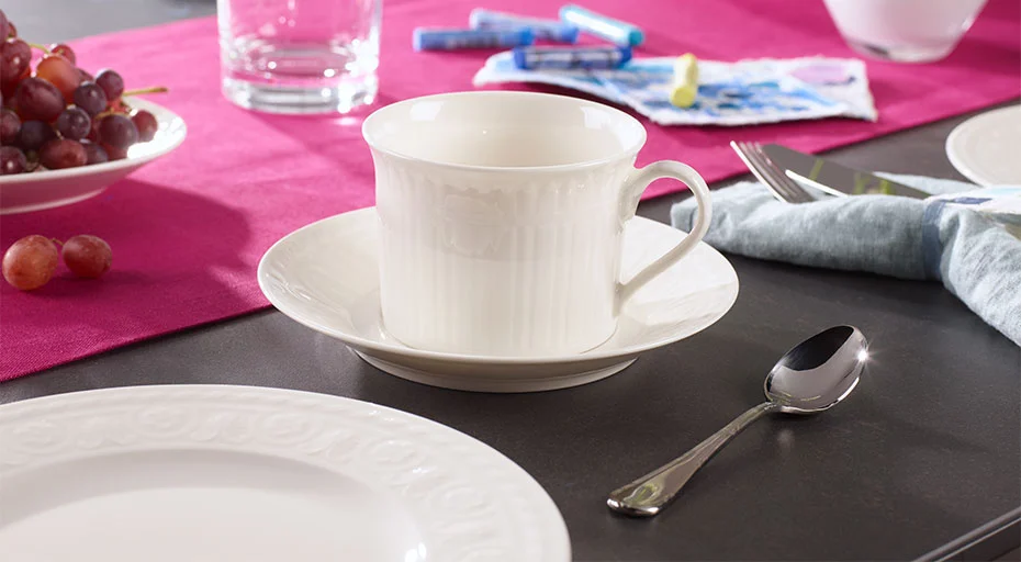 Teelöffel Kreuzband Septfontaines von Villeroy &amp; Boch neben Porzellantasse auf Untertasse auf gedecktem Tisch mit rotem Tischtuch und Trauben im Hintergrund.