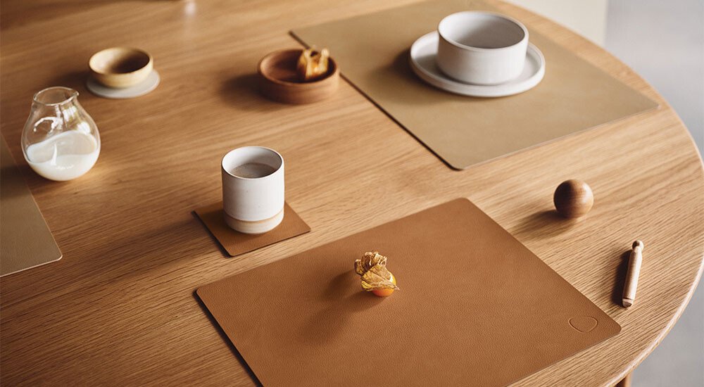 Minimalistisch gedeckter Holztisch mit Geschirr, Tassen, einer Karaffe Milch auf Tischsets der Serie Square von Lind DNA und einer Vase in warmen Naturtönen.