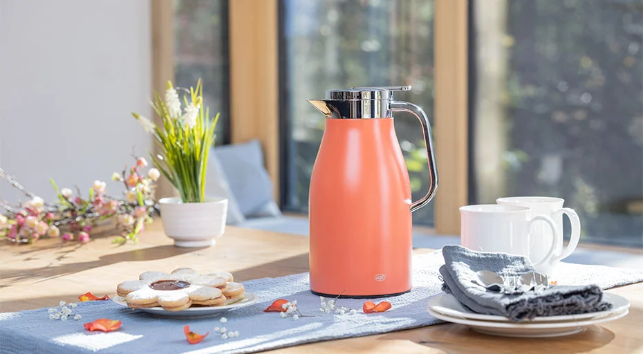 Moderne apricot-farbene Thermoskanne der Serie Skyline von Alfi auf einem Tisch mit Tassen, Keksen und Blumen in hellem, sonnigem Raum.