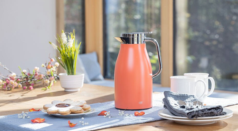 Moderne apricot-farbene Thermoskanne der Serie Skyline von Alfi auf einem Tisch mit Tassen, Keksen und Blumen in hellem, sonnigem Raum.