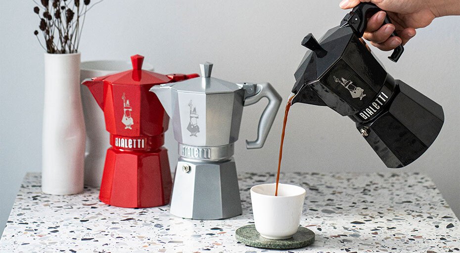 Drei Mokkakannen von Bialetti in Rot, Silber und Schwarz; eine gießt Kaffee in eine Tasse.