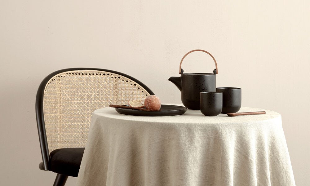 Stuhl und Tisch mit weißer Tischdecke, schwarzem Teekessel, Tassen von ASA Coppa Kuro und einem Snack auf einem Teller.