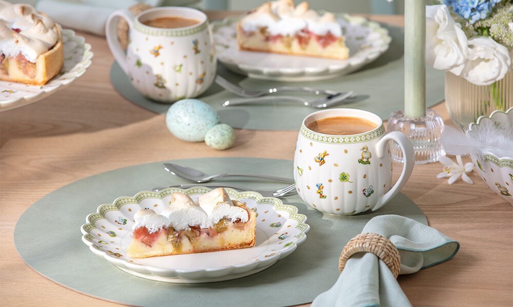 Porzellanteller der Serie Easter Delight von Villeroy &amp; Boch mit Kuchenstück und Tasse Kaffee auf gedecktem Tisch, florale Motive, Osterdeko mit Eiern und Blumen.
