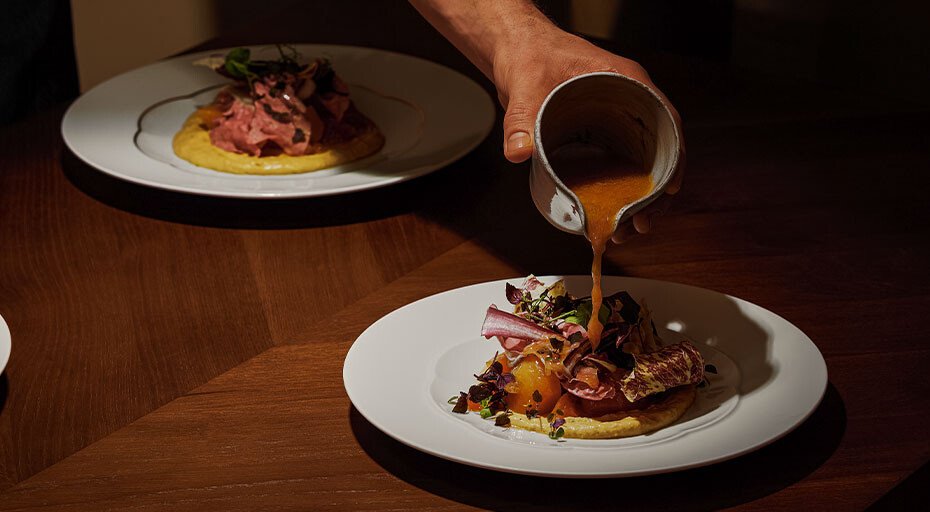 Ein Teller der Serie Sonetto Bianco von Rosenthal mit Fleisch und Gemüse wird mit einer braunen Soße aus einem Krug serviert, im Hintergrund ein weiterer Teller.