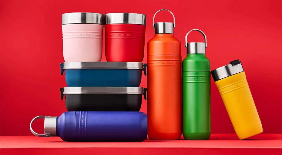 Mehrere bunte Thermosflaschen und Brotdosen der Serie On The Go von Le Creuset vor einem roten Hintergrund, gestapelt in zwei Reihen.