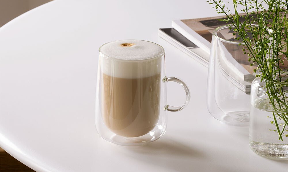 Doppelwandiges Glas der Serie Villeroy &amp; Boch Artesano Hot &amp; Cold Beverages mit Cappuccino auf weißem Tisch, im Hintergrund Bücher und eine Vase mit Pflanzen.