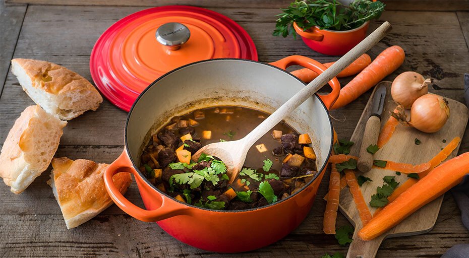Bräter von Le Creuset mit Eintopf mit Fleisch und Gemüse umgeben von Karotten, Zwiebeln, Brot und Kräutern auf Holztisch.