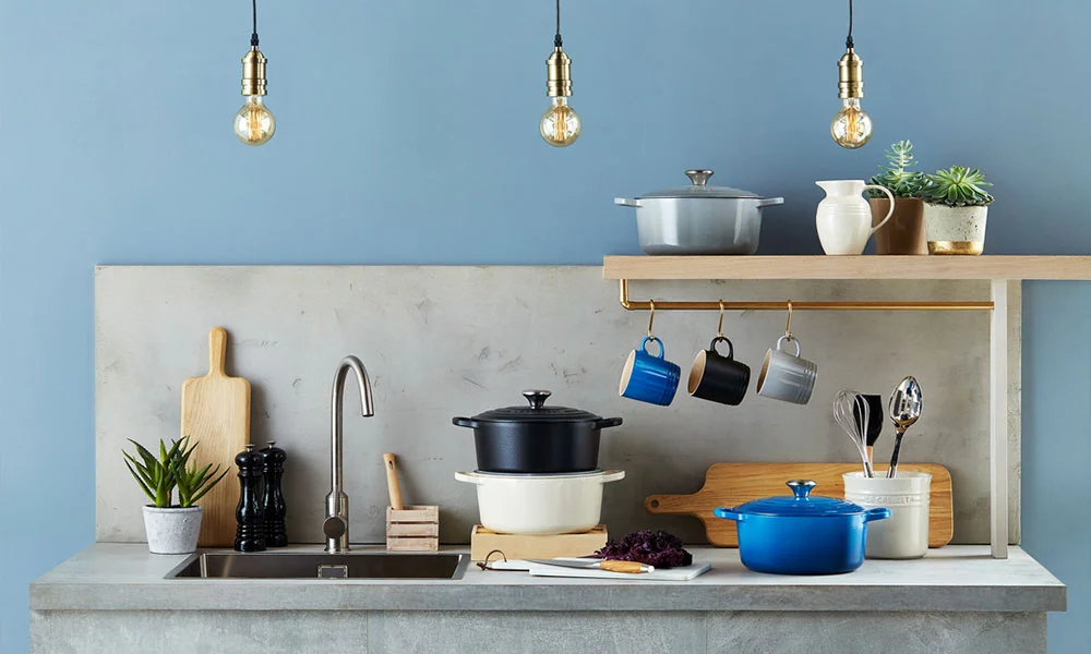 Moderne Küchenzeile mit Hängelampen, Geschirr, Pflanzen und Kochutensilien von Le Creuset auf Arbeitsplatte.