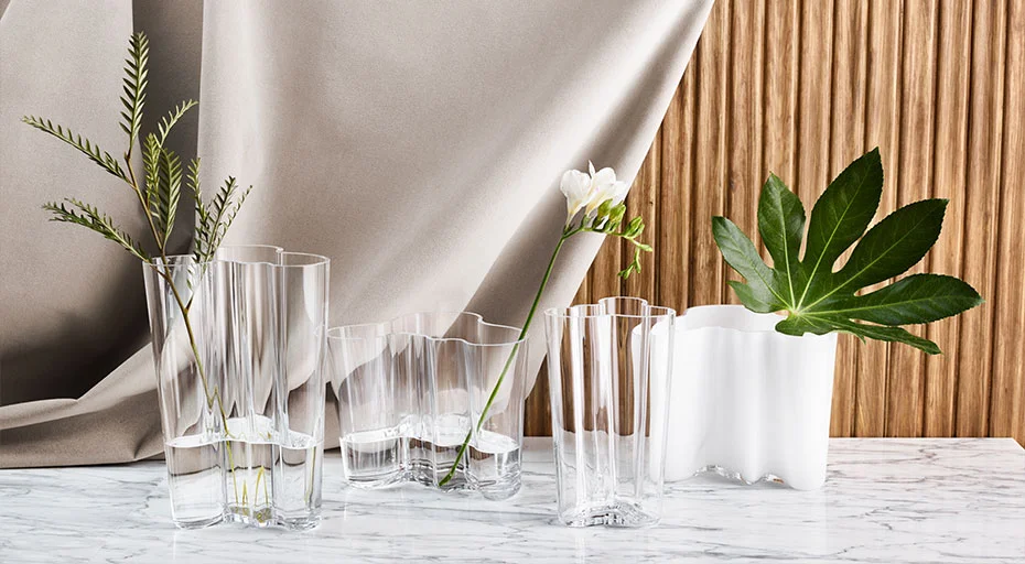 Iittala Alvar Aalto Vasen mit klaren und opaken Designs, dekoriert mit Gräsern und Blumen vor drapiertem Stoff und Holzhintergrund auf Marmortisch.