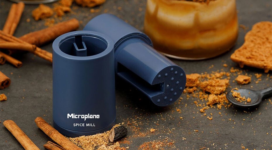 Blaue Gewürzmühle von Microplane auf Tisch mit Zimtstangen und Gewürzkrümeln, daneben ein Gewürzglas.