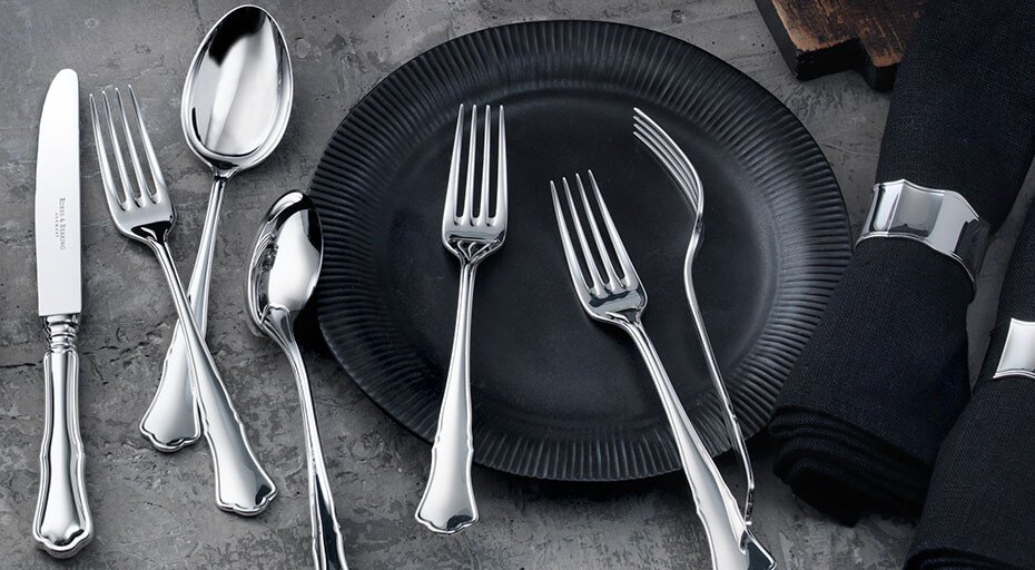 Besteck-Satz der Serie Alt Chippendale von Robbe &amp; Berking neben einem schwarzen Teller und schwarzer Serviette auf steinernem Hintergrund.
