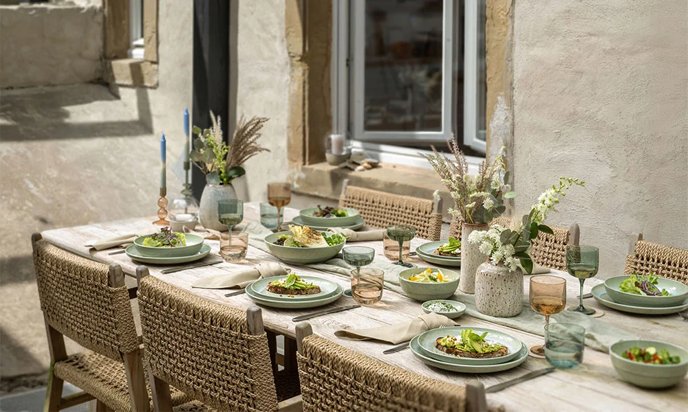 Elegante, gedeckte Tafel im Freien mit Keramikgeschirr, dekoriert mit Blumen und Kerzen, ideal für ein sommerliches Abendessen.