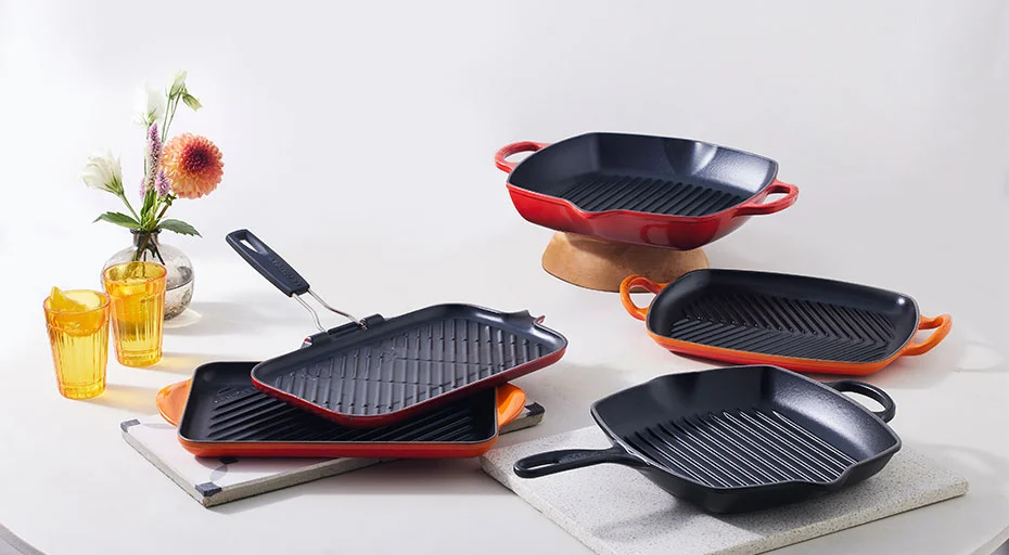 Eine Sammlung gusseiserner Bratpfannen und Grillschalen von Le Creuset in verschiedenen Größen auf einer Oberfläche mit Blumen und Gläsern.
