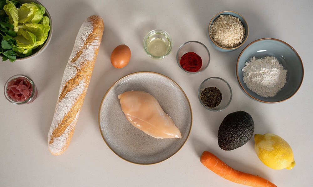 Zutaten für ein Gericht: Baguette, Hähnchenbrust, Salat, Karotte, Avocado, Zitrone, Gewürze und Öl auf einem Tisch.