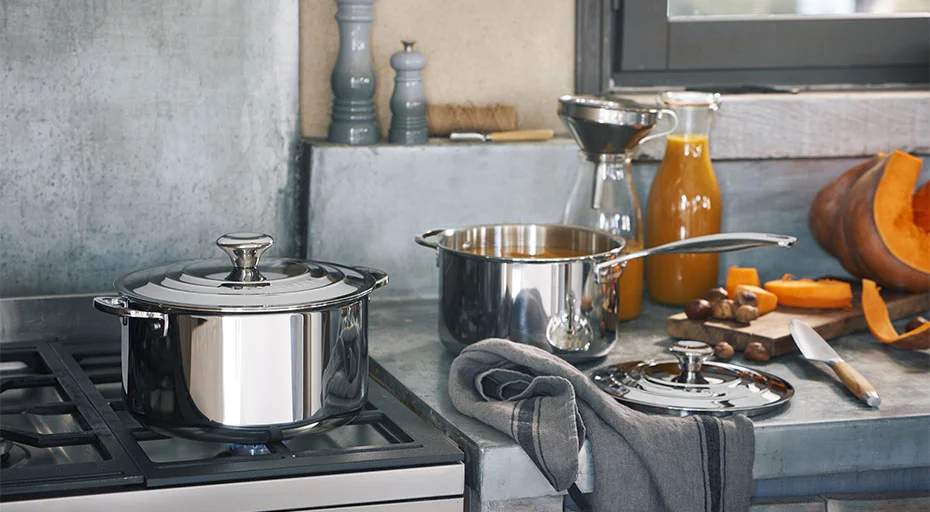 Küche mit Edelstahltöpfen der Serie 3ply von Le Creuset, Messer, Kürbis und Saft auf grauer Arbeitsplatte neben Gasherd.