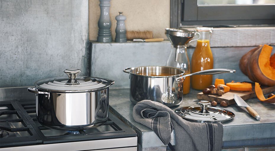 Küche mit Edelstahltöpfen der Serie 3ply von Le Creuset, Messer, Kürbis und Saft auf grauer Arbeitsplatte neben Gasherd.