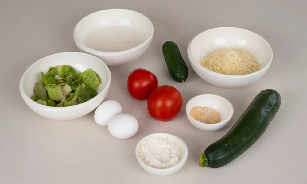 Verschiedene Zutaten in Schalen: Eier, Tomaten, Zucchini, Salat, geriebener Käse, Mehl und Gewürze auf hellem Hintergrund.
