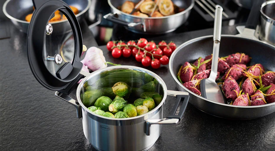 Kochtopf Silence Pro von Rösle mit Rosenkohl neben Pfanne der selben Serie auf Küchenzeile neben Tomaten und Knoblauch