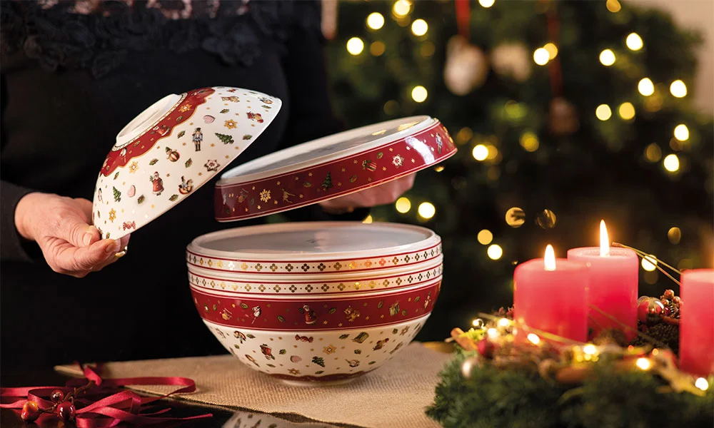 Villeroy &amp; Boch La Boule Toy's Delight Geschirr-Set, dekoriert mit Weihnachtsmotiven; im Hintergrund ein beleuchteter Weihnachtsbaum und Kerzen.