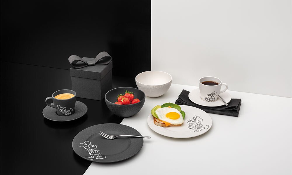 Schwarze und weiße Teller, Tassen und Schalen der Serie Villeroy &amp; Boch Manufacture Rock – Mickey Mouse, dazu Kaffee, Erdbeeren, Geschenkbox und Toast auf monochromem Hintergrund.