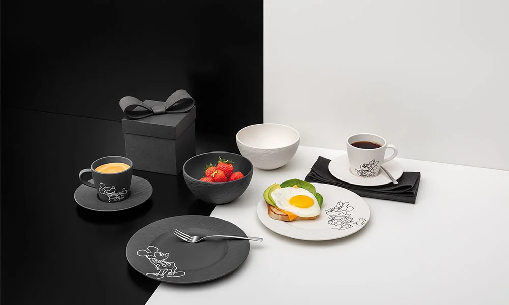 Schwarze und weiße Teller, Tassen und Schalen der Serie Villeroy &amp; Boch Manufacture Rock – Mickey Mouse, dazu Kaffee, Erdbeeren, Geschenkbox und Toast auf monochromem Hintergrund.