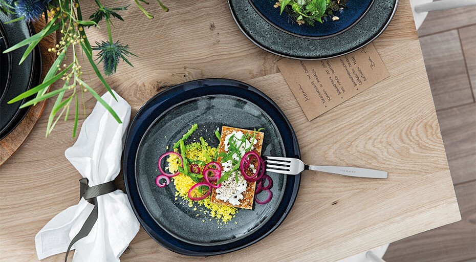 Elegant gedeckter Tisch mit kunstvoll angerichtetem Gericht auf blauer Platte, Gabel daneben, dekorative Pflanzen im Hintergrund.