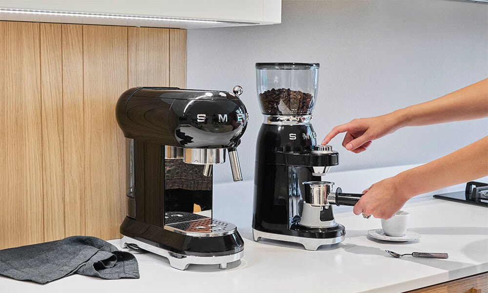 Eine schwarze Kaffeemühle und eine schwarze Espressomaschinen von Smeg auf einer weißen Küchenarbeitsplatte, menschliche Hand bedient den Mahlvorgang.