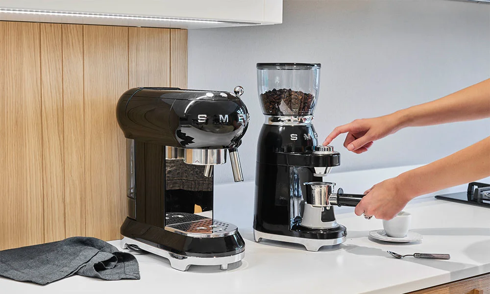 Eine schwarze Kaffeemühle und eine schwarze Espressomaschinen von Smeg auf einer weißen Küchenarbeitsplatte, menschliche Hand bedient den Mahlvorgang.