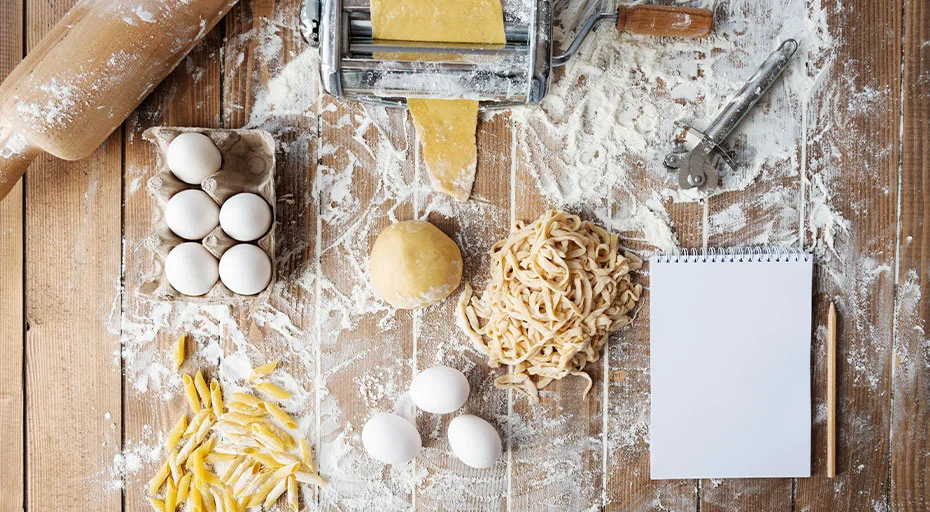 Teigwarenherstellung: Nudelmaschine, Teig, Eier, hartes und gerolltes Mehl auf Holztisch, Notizblock zur Rezeptaufzeichnung.
