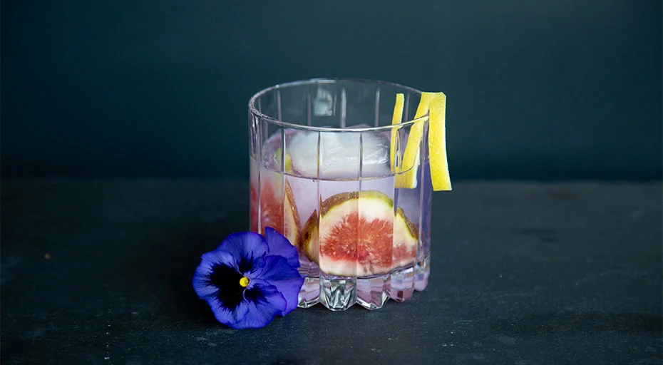 Ein Glas der Serie Resort von Riedel mit Cocktail, Eiswürfeln, Feigenscheiben und Zitronenzeste, dekoriert mit einer violetten Blüte, vor dunklem Hintergrund.