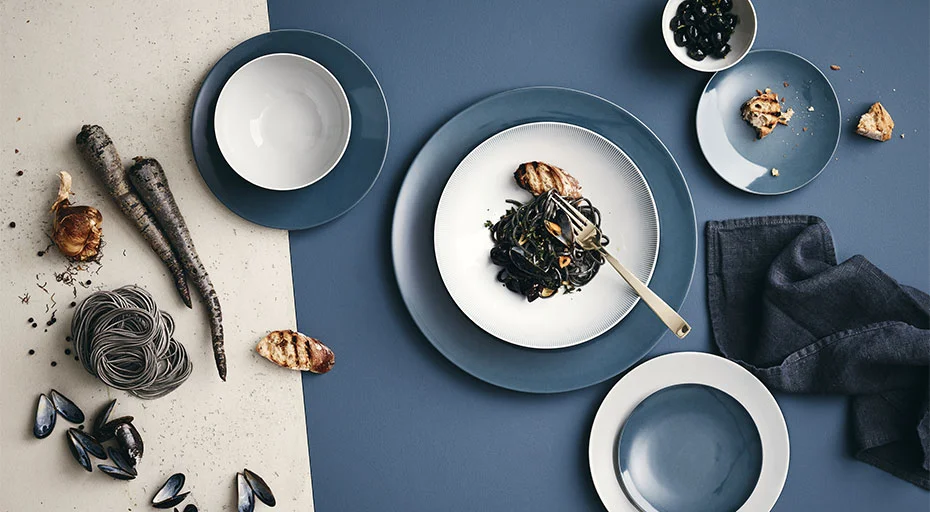 Rosenthal TAC auf elegantem Tischarrangement mit blauen Tellern, Sepia-Nudeln, Grissini, Muscheln und dekorativen Karotten.