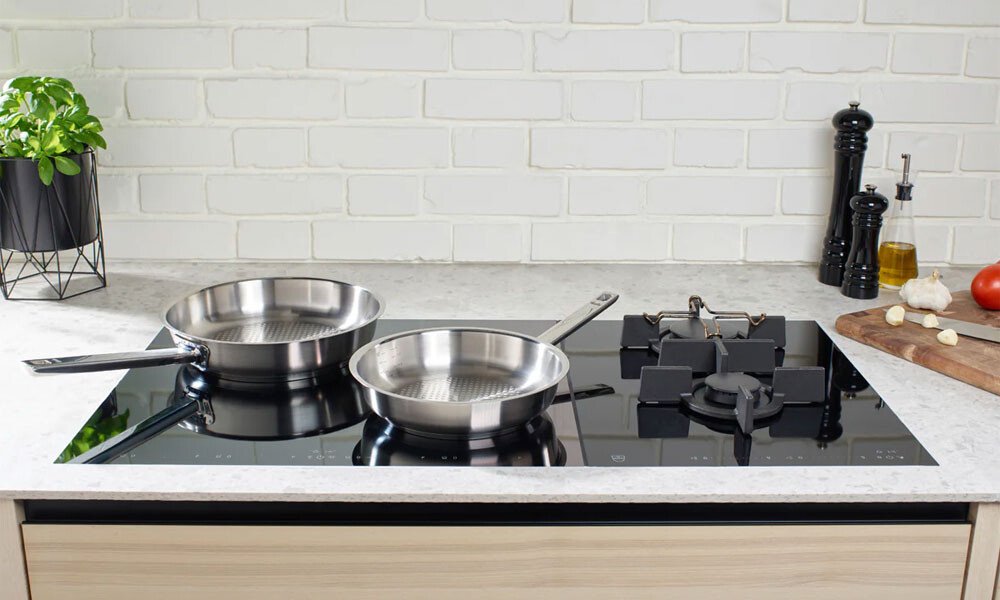 Zwei Edelstahlpfannen der Original Profi Collection von Fissler auf einem modernen Induktionskochfeld, umgeben von Gewürzen und frischen Zutaten.