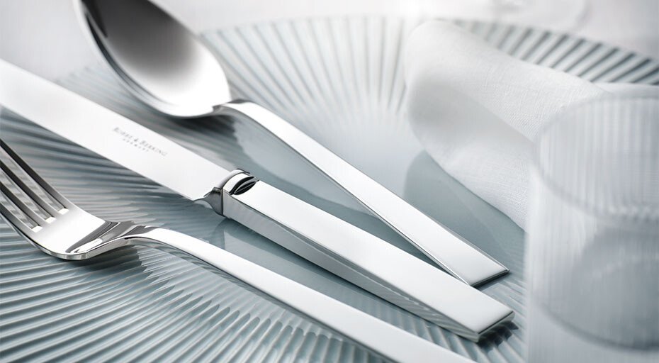 Besteck der Serie Riva von Robbe &amp; Berking auf einem strukturierten Glasteller, in stilvoller Anordnung mit Glas und Serviette.