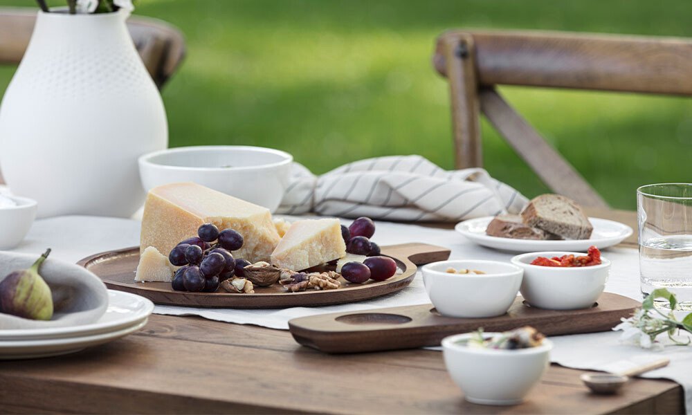Holztisch gedeckt mit Käseplatte, Trauben, Nüssen, Brot und Geschirr der Serie Artesano von Villeroy &amp; Boch im Freien. Hintergrund: unscharfer grüner Rasen.