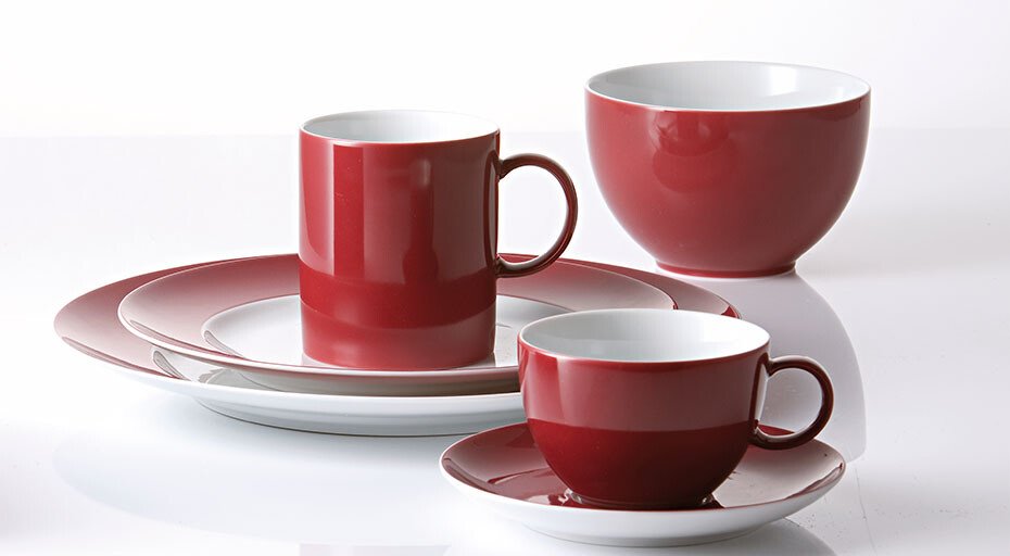 Rotes Geschirrset der Serie Sunny Day Fuchsia von Thomas, bestehend aus Tassen, Untertassen, Tellern und einem Schüssel auf einer glänzenden Oberfläche.