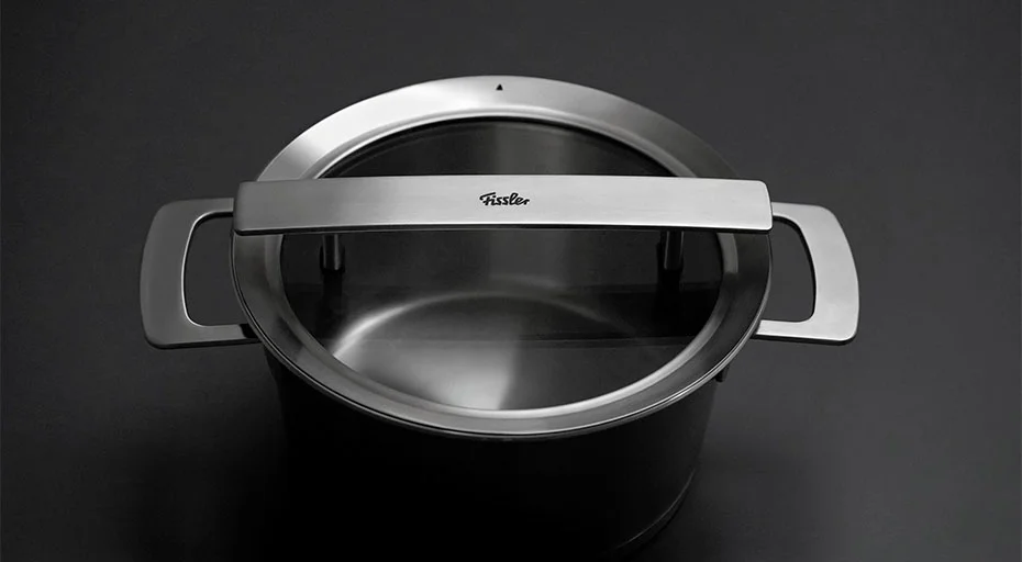 Fissler Edelstahlkochtopf mit Glasdeckel und Metallgriffen auf grauem Hintergrund.