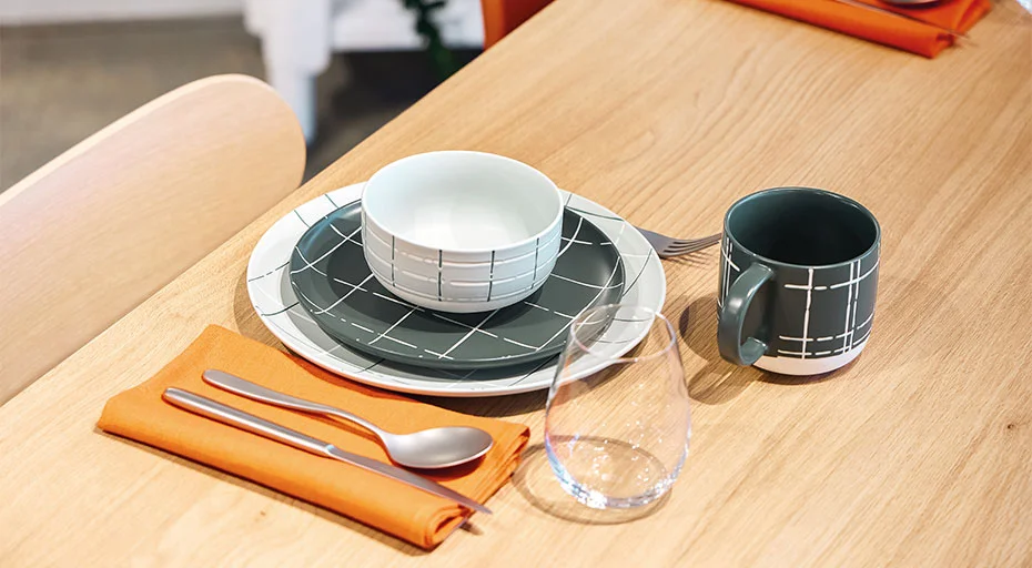 Gedeckter Holztisch mit Teller, Schüssel, Tasse der Serie FUN Karo von Thomas in geometrischem Muster, samt Besteck, Glas und orange Serviette.