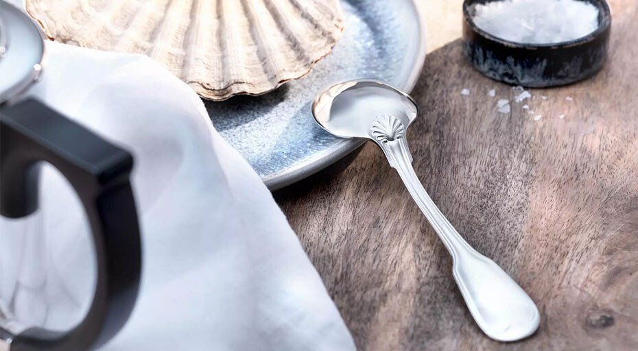 Silberlöffel der Kollektion Alt Faden von Robbe &amp; Berking auf Holztisch neben Muschel und Glasbehälter mit grobem Salz. Elegante, nautische Tischdekoration.