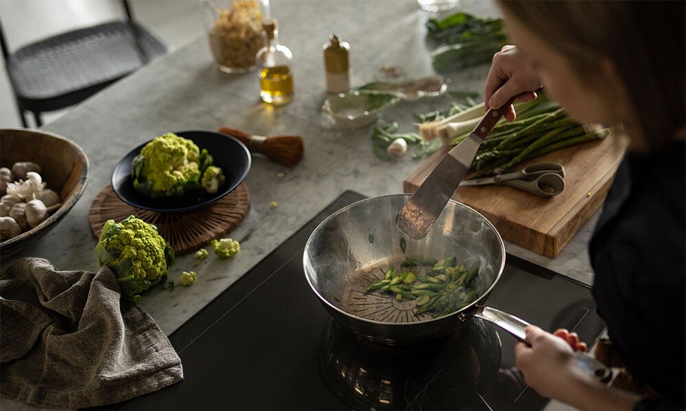 Person brät grünen Spargel in einer Pfanne von Fiskars auf einem Kochfeld. Frische Zutaten und Gewürze liegen auf dem Tisch bereit.