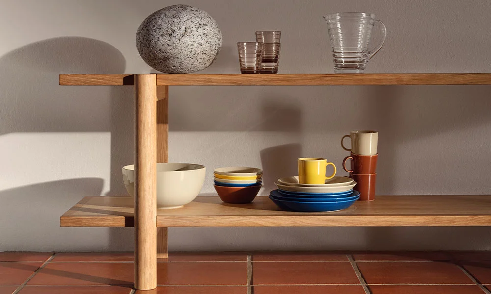 Iittala Teema Geschirr in Holzregal mit Keramikobjekten, Gläsern und Geschirr, auf gefliesten Boden, seitlich beleuchtet.