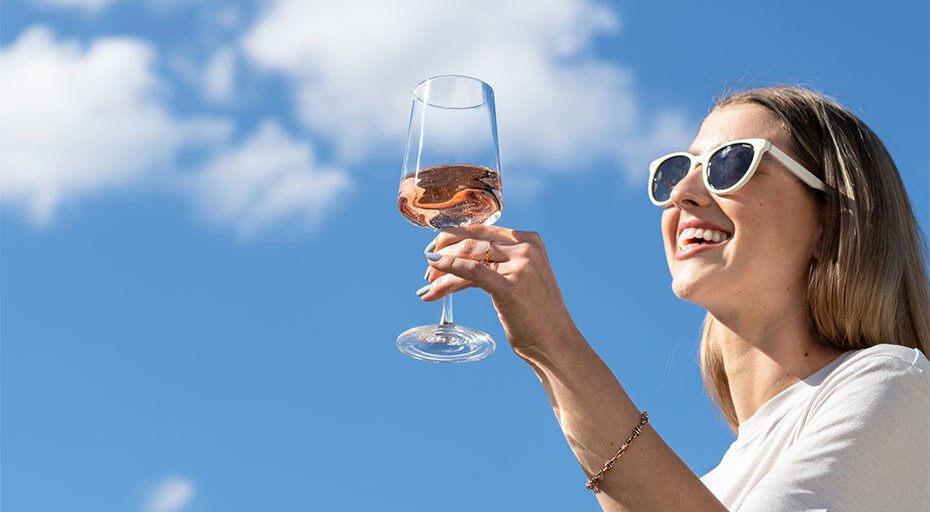 Frau im Sonnenlicht hält ein Glas der Serie MioVino von Schott Zwiesel, befüllt mit Roséwein vor blauem Himmel und Wolken, trägt Sonnenbrille und lächelt.