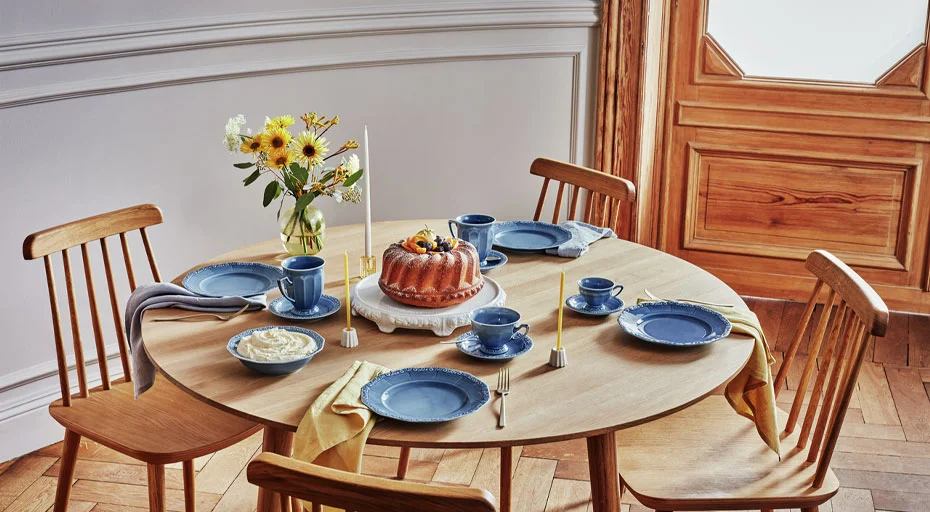 Runder Holztisch mit blauem Geschirr der Serie Maria Dream Blue von Rosenthal, Gugelhupf, Kerzen, Blumenstrauß und vier Stühlen in elegantem Interieur.