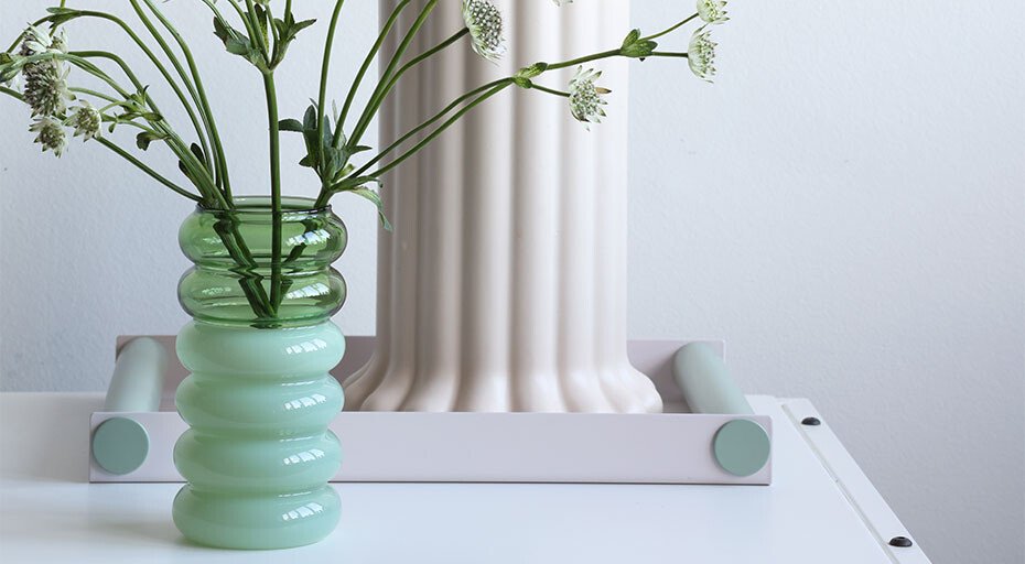 Grüne Glasvase von Design Letters mit weißen Blumen vor pastellfarbenem Säulenobjekt auf weißem Untergrund.