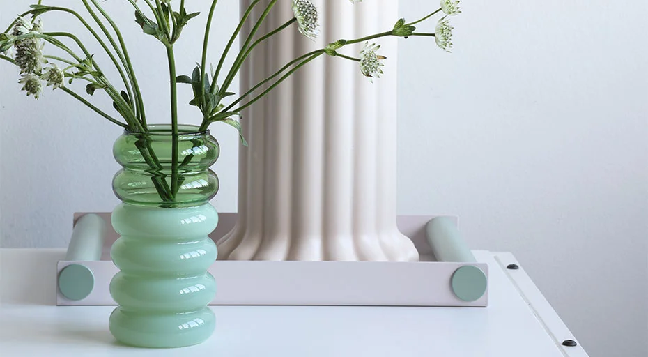 Grüne Glasvase von Design Letters mit weißen Blumen vor pastellfarbenem Säulenobjekt auf weißem Untergrund.