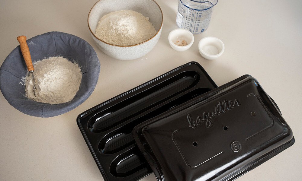 Teigzutaten und Baguette-Backform auf Tisch: Mehl in Schüssel und Tuch, Wasser, Salz, Hefe, schwarze Baguetteform.