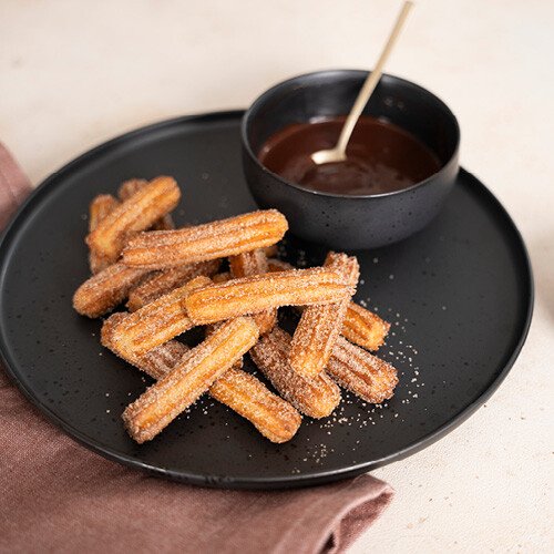 Churros mit Zimt und Zucker auf schwarzem Teller, daneben Schälchen mit Schokoladensauce und goldenem Löffel.
