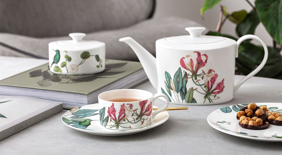Teekanne, Tasse und Untertasse der Avarua-Serie von Villeroy &amp; Boch mit Blumenmuster auf einem Tisch, daneben ein Teller mit Nüssen und ein geschlossenes Gefäß.